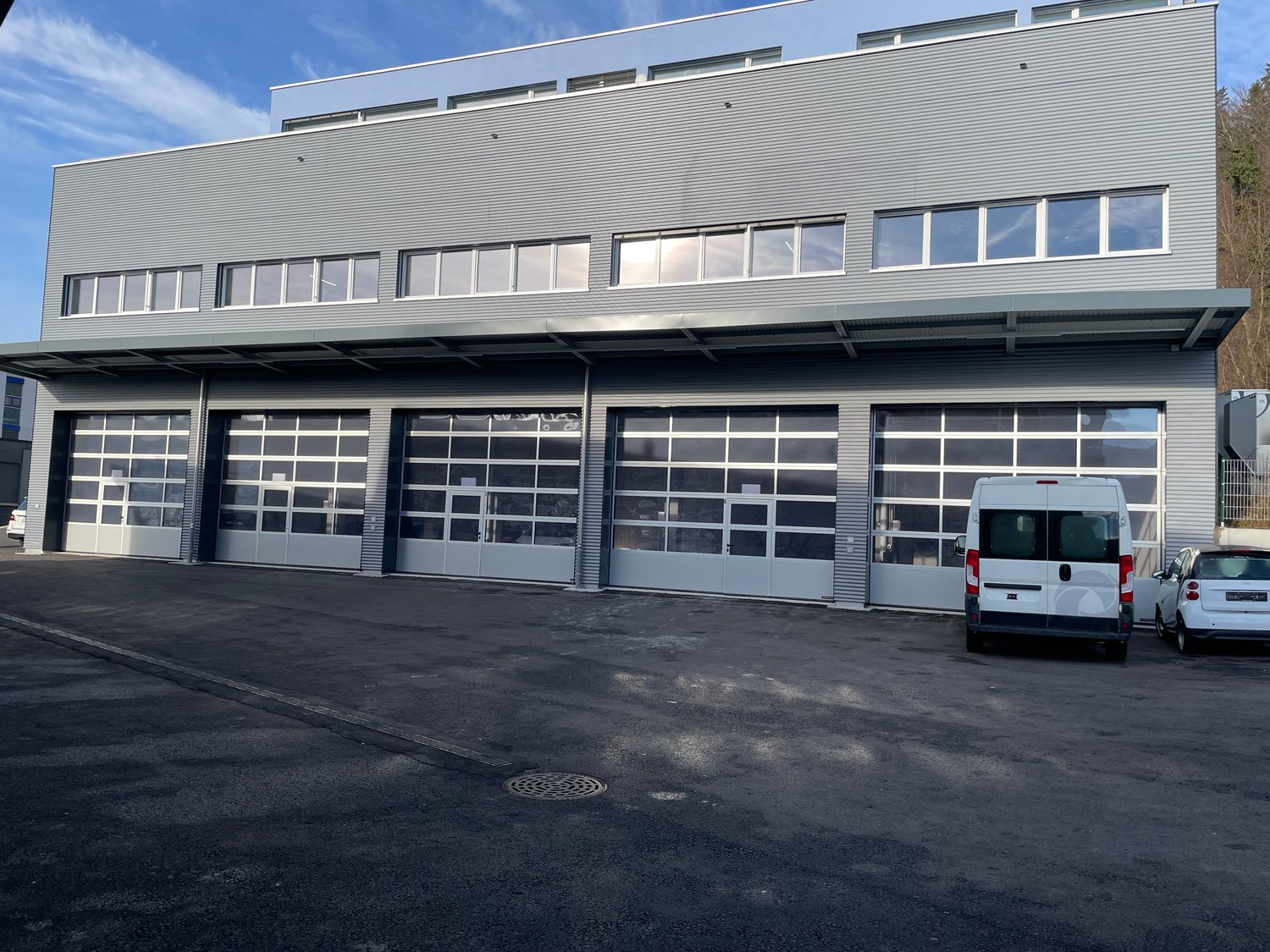modern industrial building, gray exterior, large glass doors, multiple windows, parking area