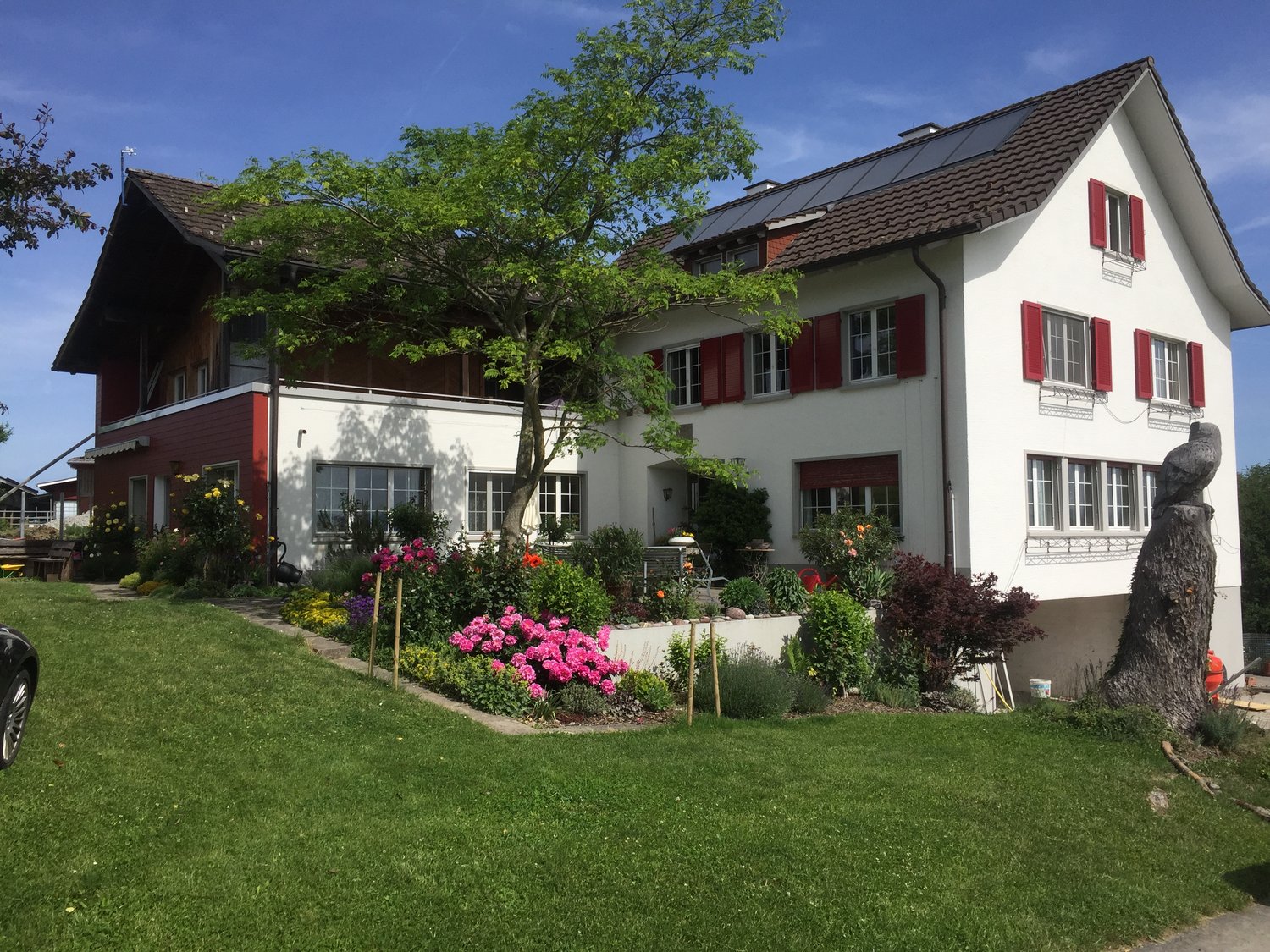 2 story house, white exterior, red shutters, solar panels on roof, red brick facade, well-maintained garden with various flowers, car parked on the side