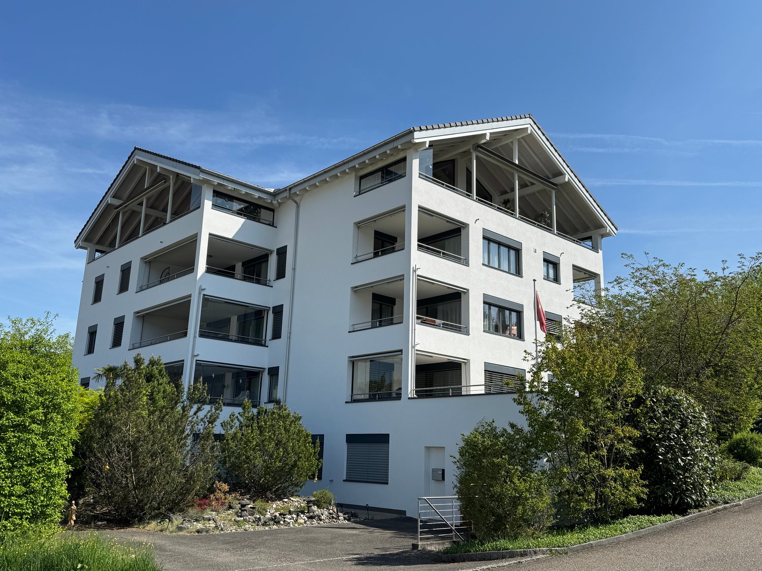 4-story apartment building with white exterior, balconies, and large windows. The building is surrounded by trees and greenery.