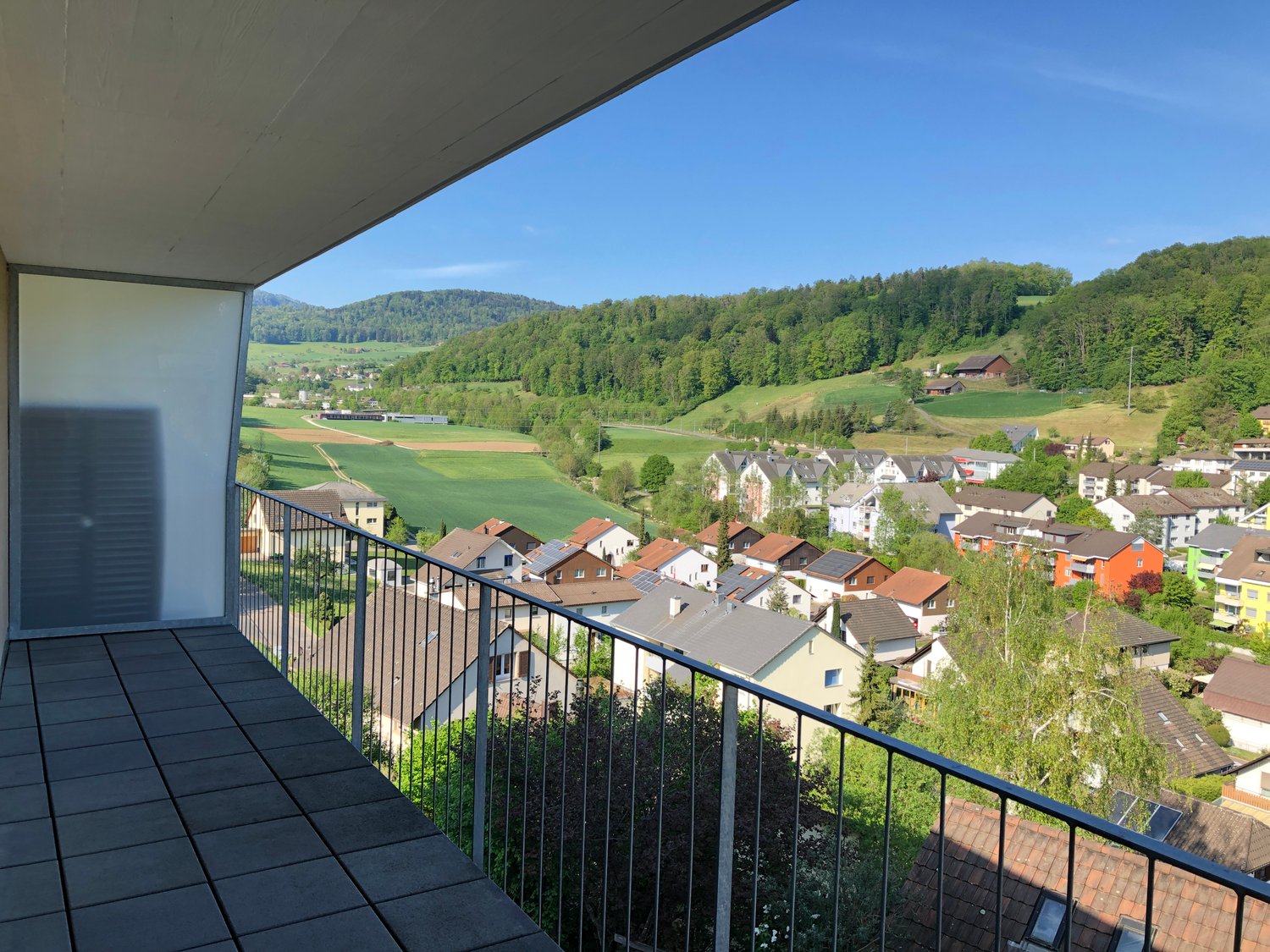 An elevated house with a balcony offering a scenic view of a countryside landscape with several buildings, hills, and a blue sky.