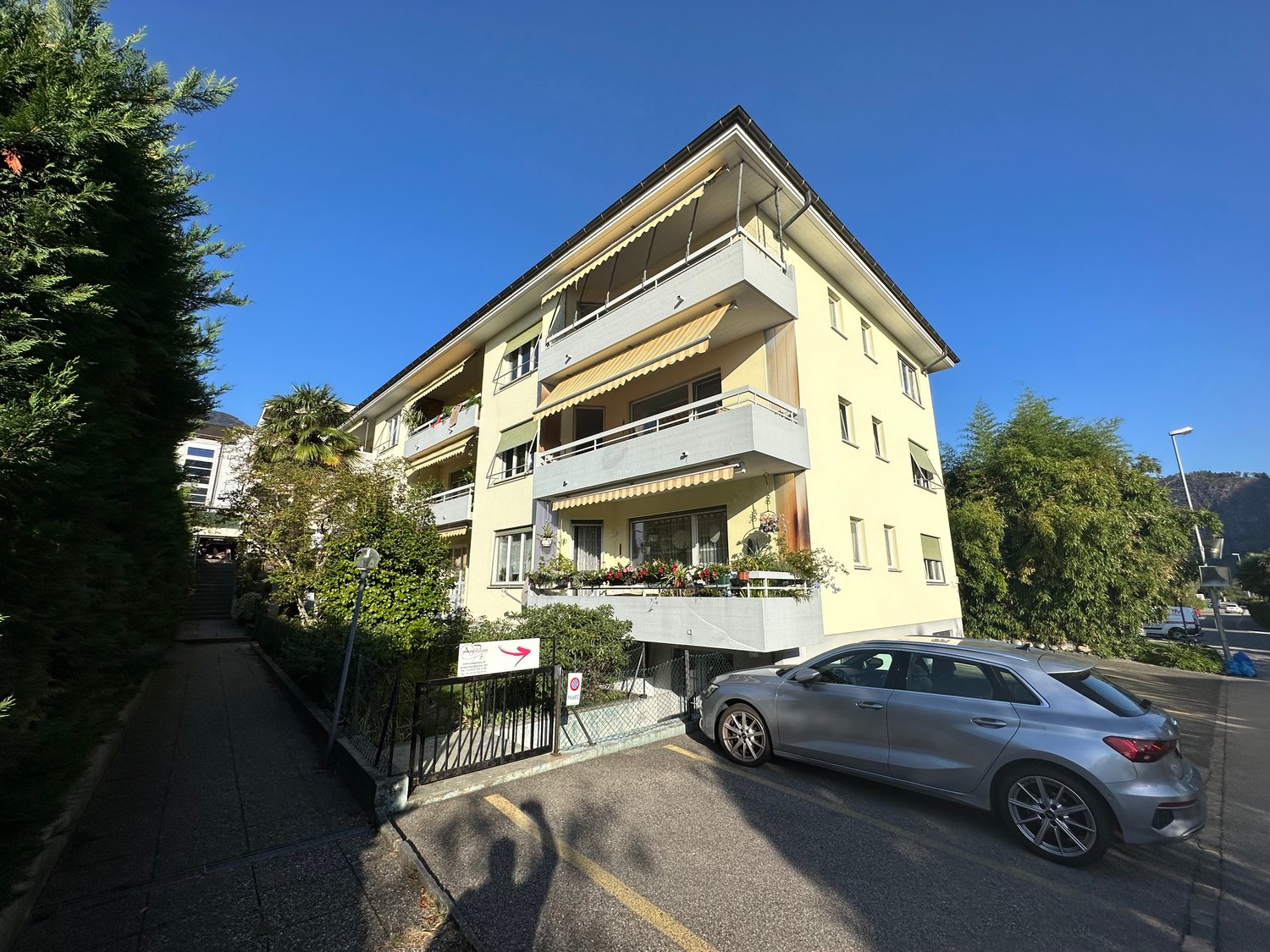 3-story yellow apartment building with balconies, surrounded by trees and vegetation, with a parked car in the foreground