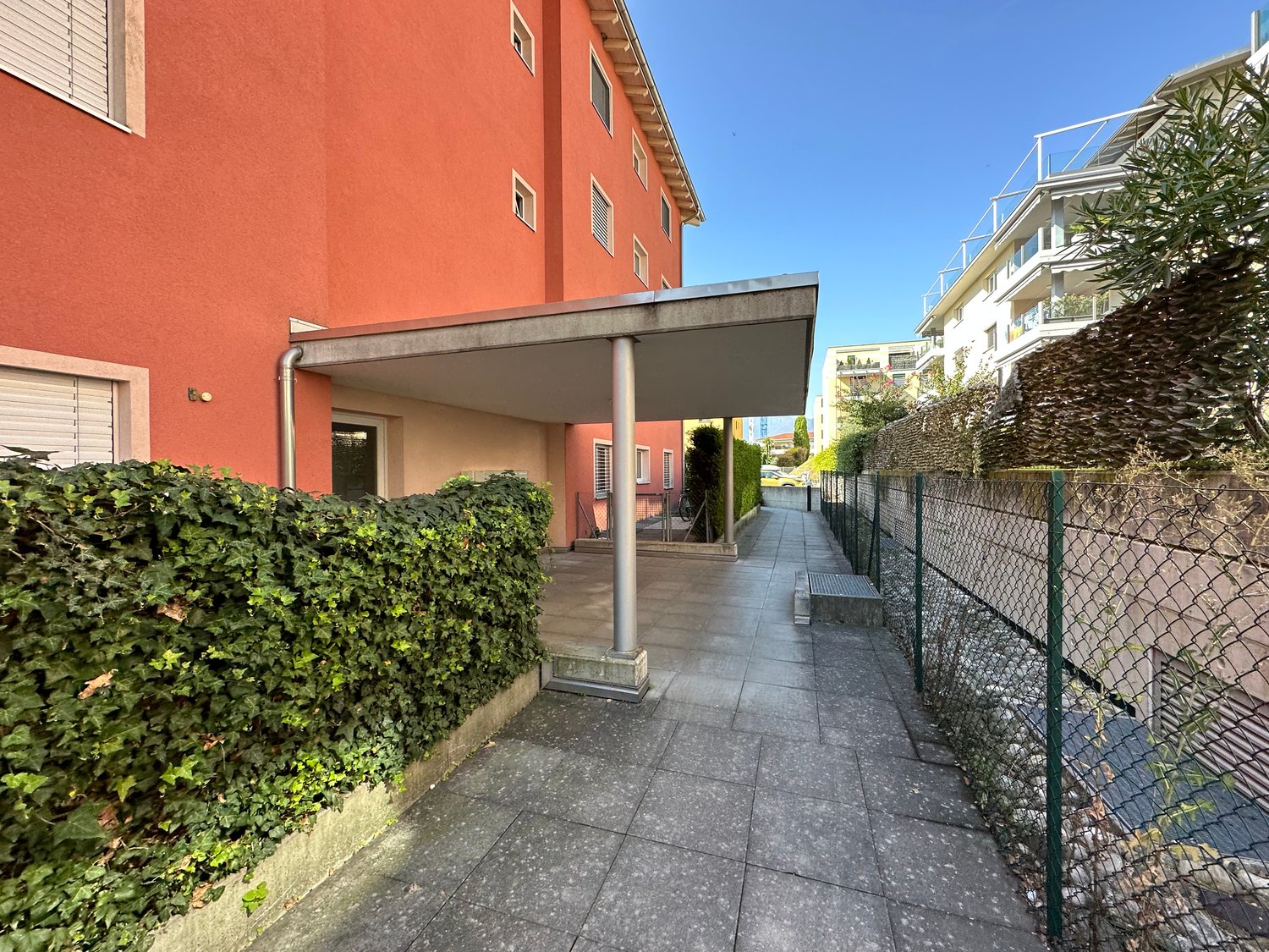 Main entrance covered by a canopy, orange colored building, large greenery bush, pathway leading to the entrance, chain link fence on the side