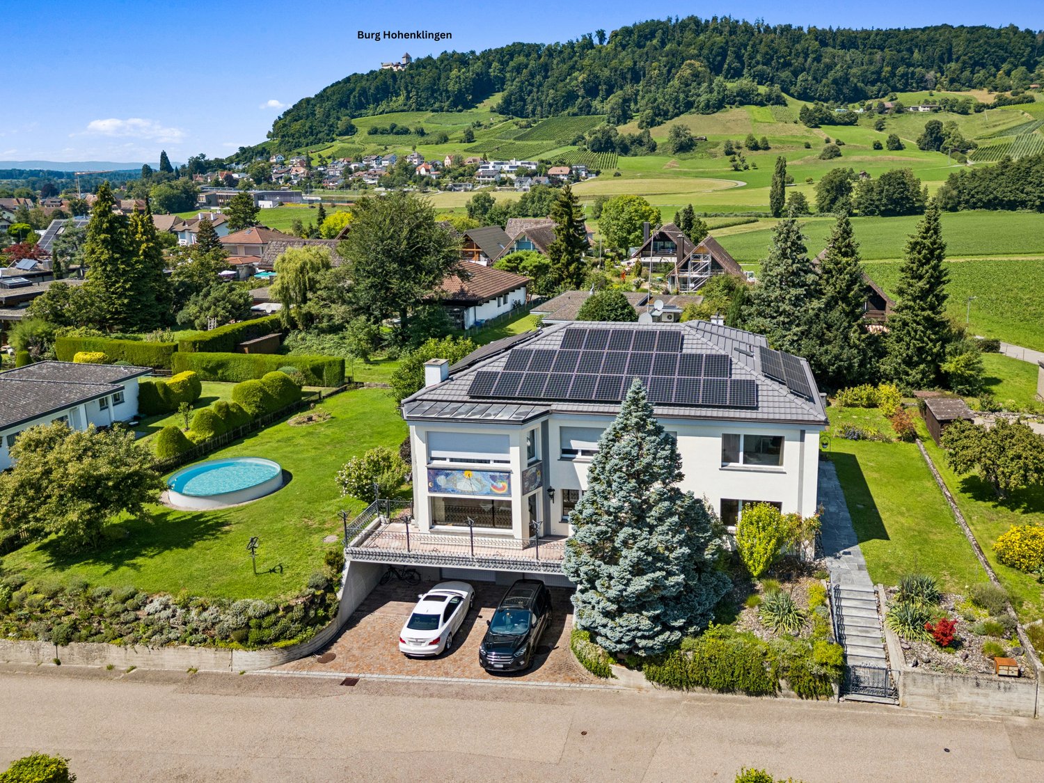 Modern white house with solar panels on roof, large outdoor swimming pool, garden, 2 cars parked in garage
