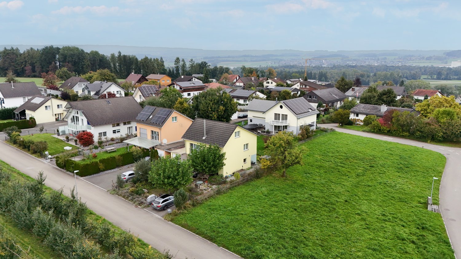 A neighborhood consisting of various single family houses with well-maintained lawns and trees. Several houses have solar panels on their roofs. The neighborhood is surrounded by green fields and a mountain range.