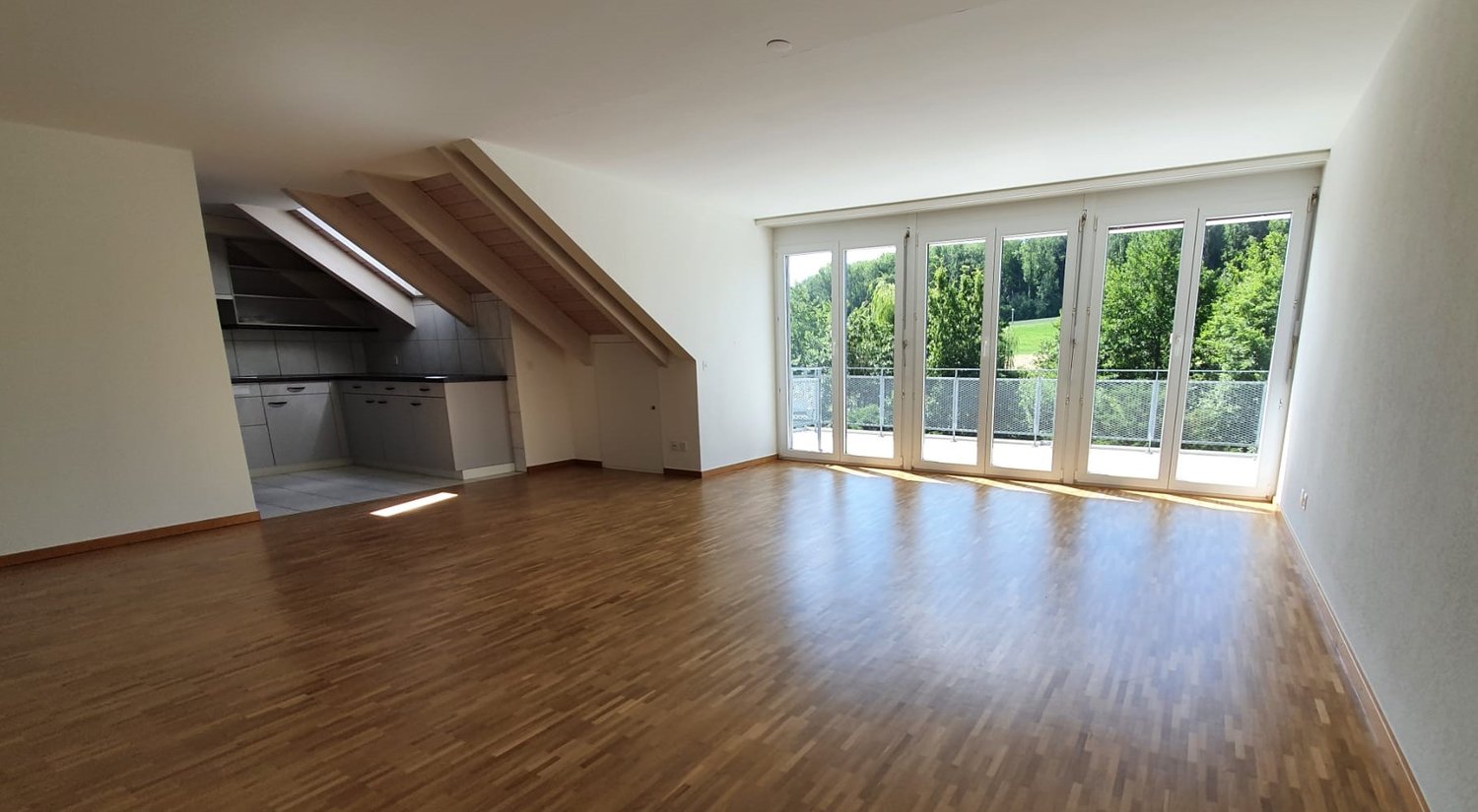 Spacious open-plan living area with hardwood flooring, large windows providing ample natural light, and access to a balcony or terrace overlooking a green outdoor area. The kitchen is integrated into the living space and features modern appliances.