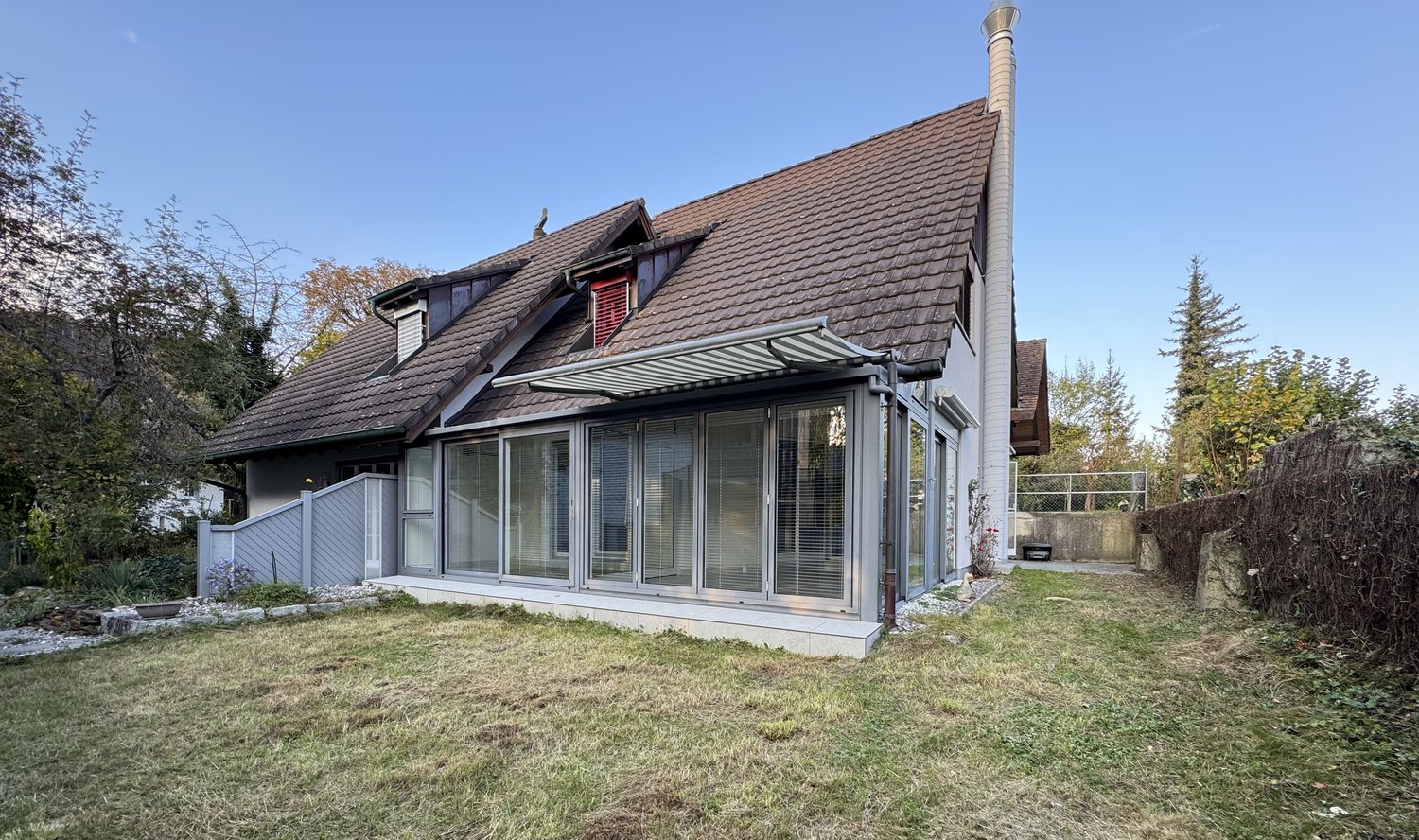 Single-family house with brown roof, glass windows, concrete raised ground floor, canopy, backyard with grass, and an extension at the side.