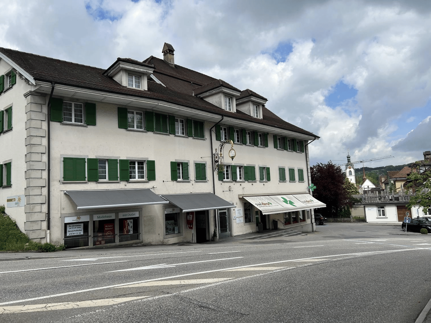 Building with shops on the ground floor, multiple windows with green shutters, chimney, awnings over windows, street sign, parked car