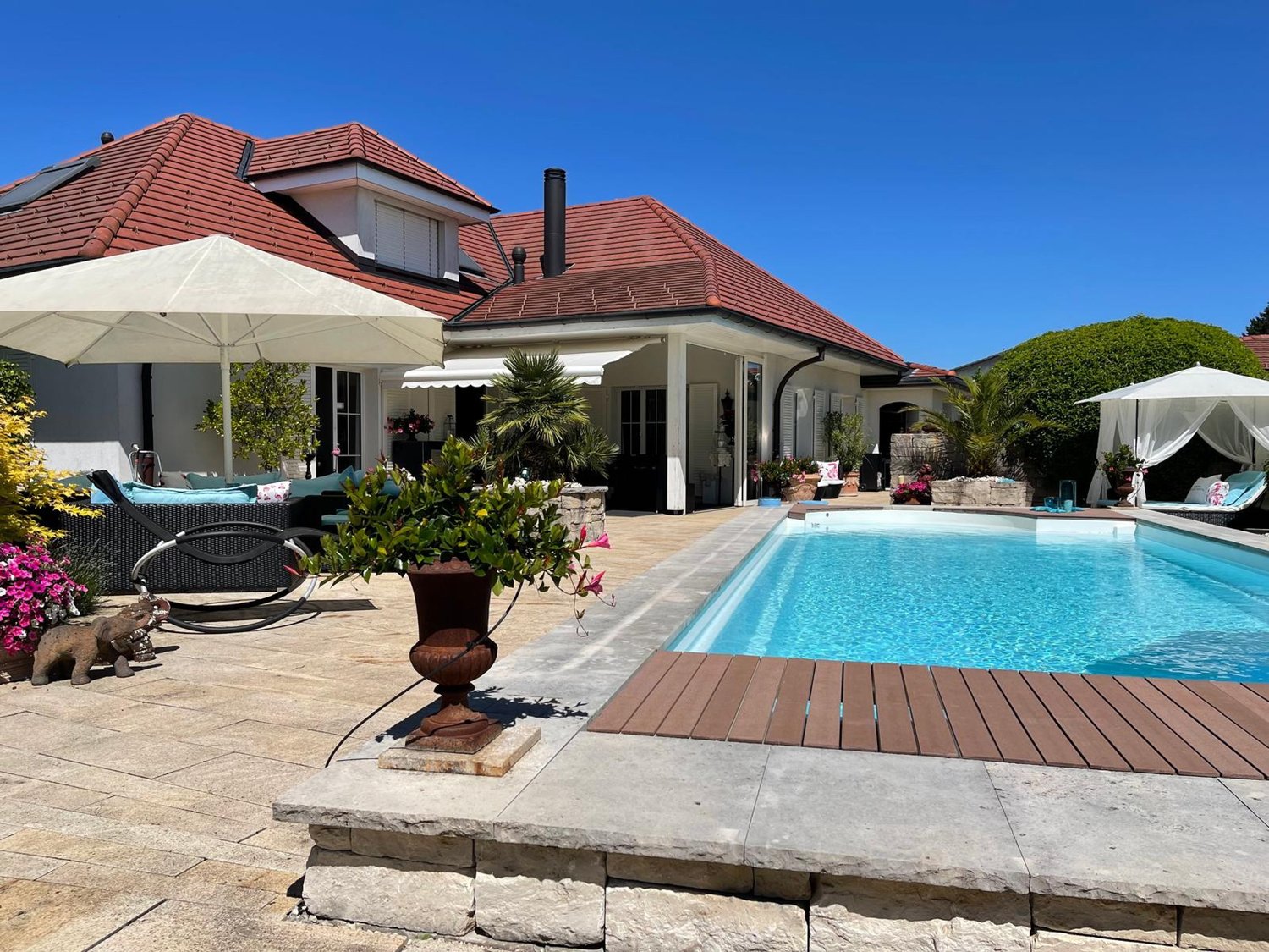 Swimming pool with wooden deck, surrounded by brick paving, patio with lounge chairs, and a large patio umbrella. There are also plants and flowers, and a wooden lounge chair with a canopy on the right side.