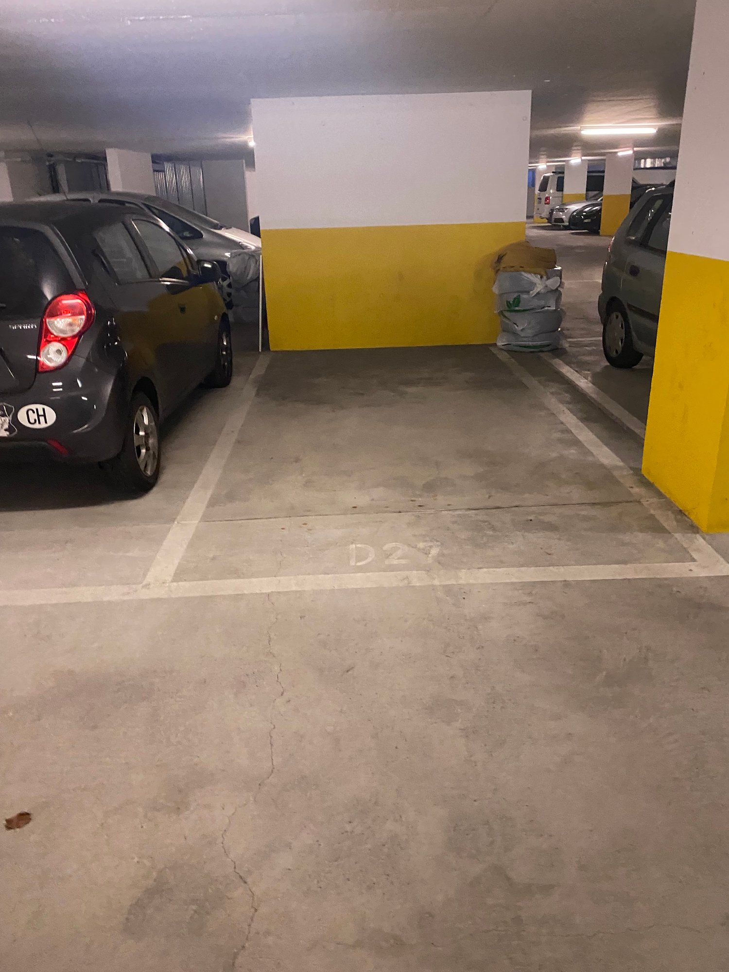 Underground parking with two available parking spaces labeled D21, concrete flooring, a yellow and white wall, a trash can near the wall, and cars parked inside.
