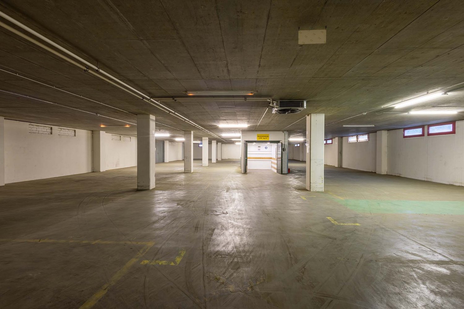 Underground parking lot, concrete floor, white walls, ceiling lights, yellow parking lines, entrance on the right