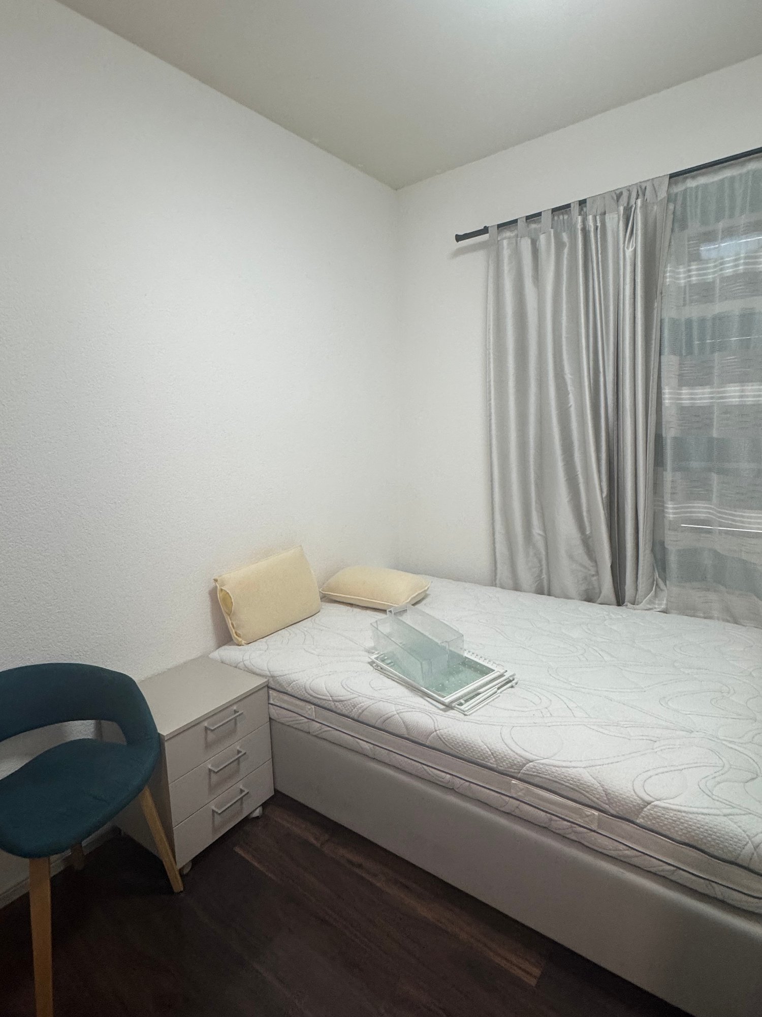 single bed, two pillows, a chair, a small table with drawers, grey curtains, wooden floor