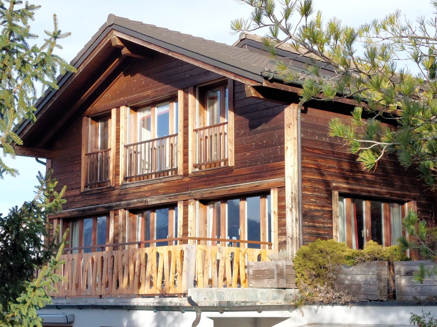 2 story wooden chalet, brown shingles, multiple glass windows, balcony with wooden railings, green trees in front
